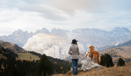 Frau steht mit ihrem Hund auf einem Felsen und blickt auf die Dolomiten – Herbstlandschaft in Südtirol mit buntem Bergwald.