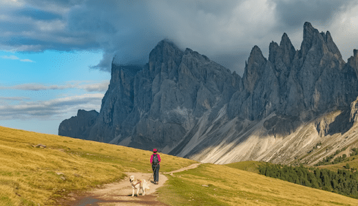 Wanderin mit Hund auf einem Bergpfad in den Dolomiten, im Hintergrund beeindruckende Felsgipfel und ziehende Wolken.