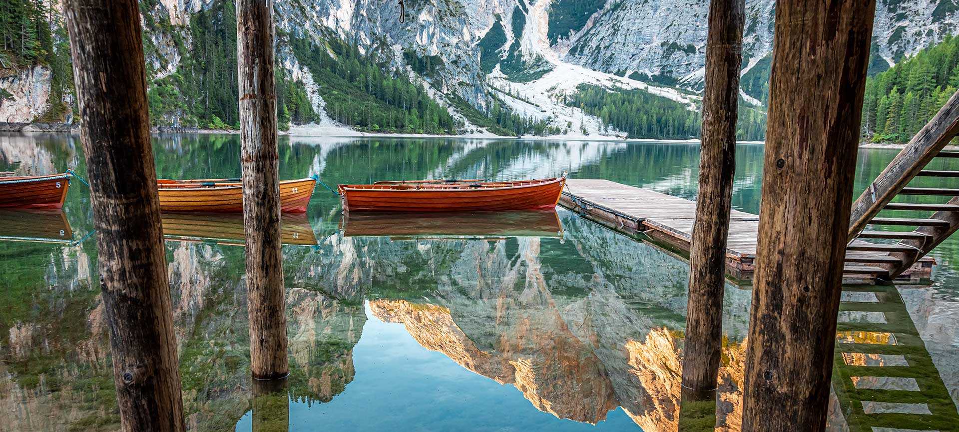Blick vom Bootshaus auf den Pragser Wildsee Blick durch die Holzpfähle des historischen Bootshauses am Pragser Wildsee auf aufgereihte Ruderboote und die Bergspiegelung